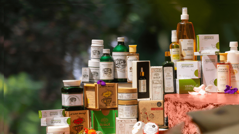 A premium display of Kairali Ayurvedic products including herbal oils, skincare, wellness supplements, and Ayurvedic medicine arranged elegantly with natural floral accents against a green nature-inspired background.
