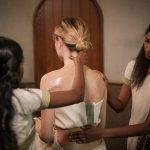 Two Ayurvedic therapists apply an oil massage to a seated woman’s back and neck in a spa treatment room.
