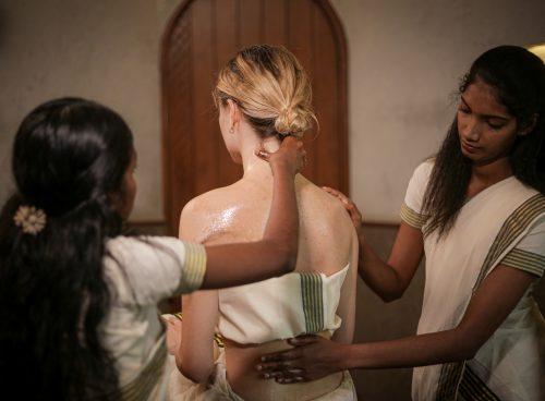 Two Ayurvedic therapists apply an oil massage to a seated woman’s back and neck in a spa treatment room.