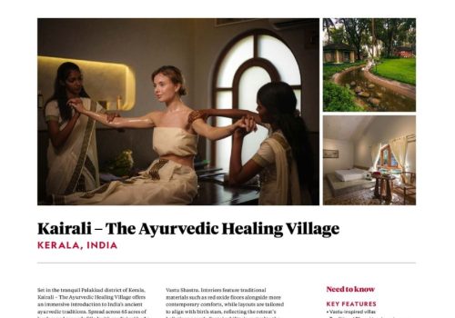 Two Ayurvedic therapists in traditional white sarees performing a synchronised arm massage treatment on a female guest at Kairali – The Ayurvedic Healing Village in Kerala, India, inside a warmly lit treatment room with an arched window.