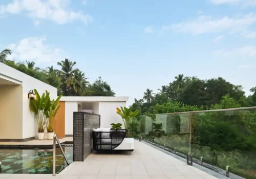 Modern outdoor poolside terrace with a lounge chair, glass railing, and tropical greenery under a blue sky.