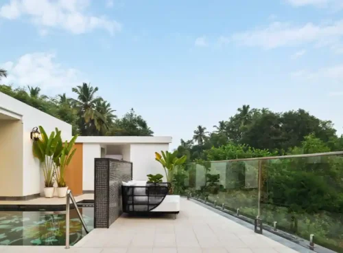 Modern outdoor poolside terrace with a lounge chair, glass railing, and tropical greenery under a blue sky.