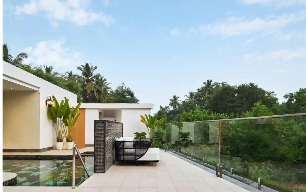 Modern outdoor poolside terrace with a lounge chair, glass railing, and tropical greenery under a blue sky.