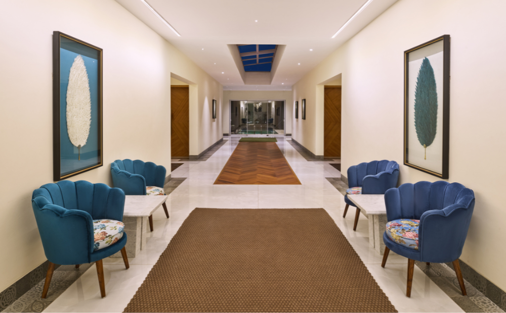 Elegant hotel corridor with blue velvet scallop chairs, botanical artwork, herringbone wood floor inlay, and a skylight at a luxury property in Goa