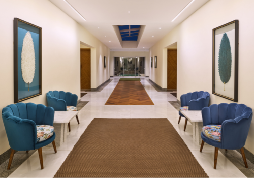 Elegant hotel corridor with blue velvet scallop chairs, botanical artwork, herringbone wood floor inlay, and a skylight at a luxury property in Goa
