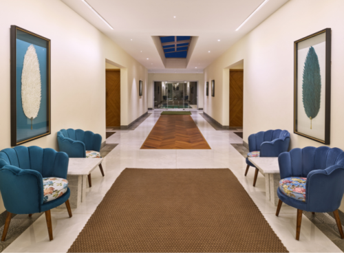 Elegant hotel corridor with blue velvet scallop chairs, botanical artwork, herringbone wood floor inlay, and a skylight at a luxury property in Goa