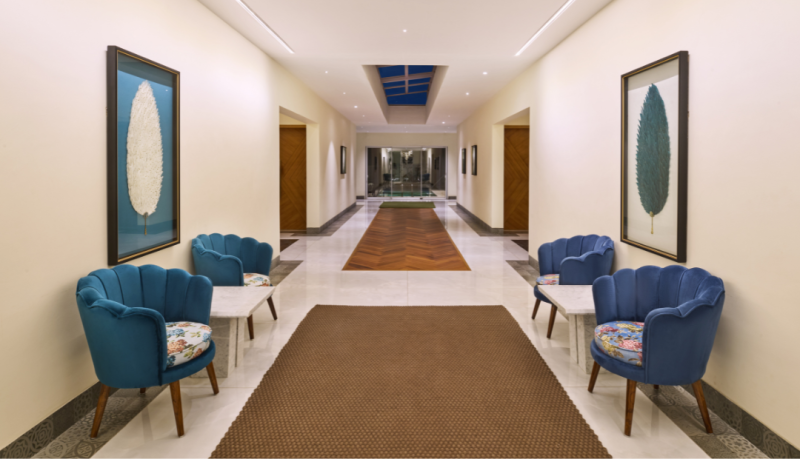 Elegant hotel corridor with blue velvet scallop chairs, botanical artwork, herringbone wood floor inlay, and a skylight at a luxury property in Goa