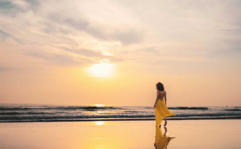 Woman in yellow dress walking alone on beach at sunset, embracing slow travel