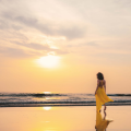Woman in yellow dress walking alone on beach at sunset, embracing slow travel