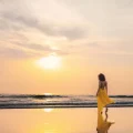 A woman in a flowing yellow dress walks along a calm beach at golden hour, her reflection mirrored in the wet sand. The soft pastel sky and gentle waves capture the essence of slow travel — taking time to be present, unhurried, and fully immersed in the beauty of a destination rather than rushing through it.