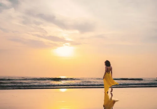 A woman in a flowing yellow dress walks along a calm beach at golden hour, her reflection mirrored in the wet sand. The soft pastel sky and gentle waves capture the essence of slow travel — taking time to be present, unhurried, and fully immersed in the beauty of a destination rather than rushing through it.