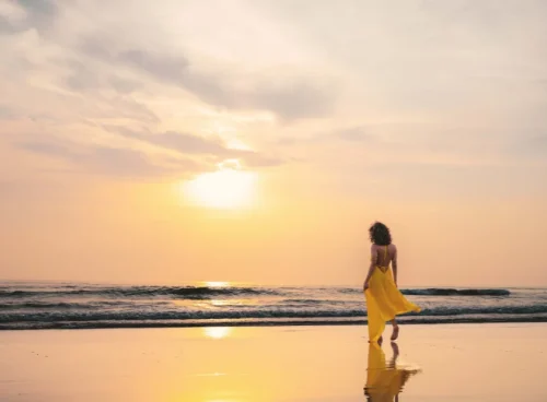 A woman in a flowing yellow dress walks along a calm beach at golden hour, her reflection mirrored in the wet sand. The soft pastel sky and gentle waves capture the essence of slow travel — taking time to be present, unhurried, and fully immersed in the beauty of a destination rather than rushing through it.