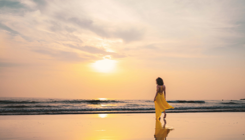 Woman in yellow dress walking alone on beach at sunset, embracing slow travel