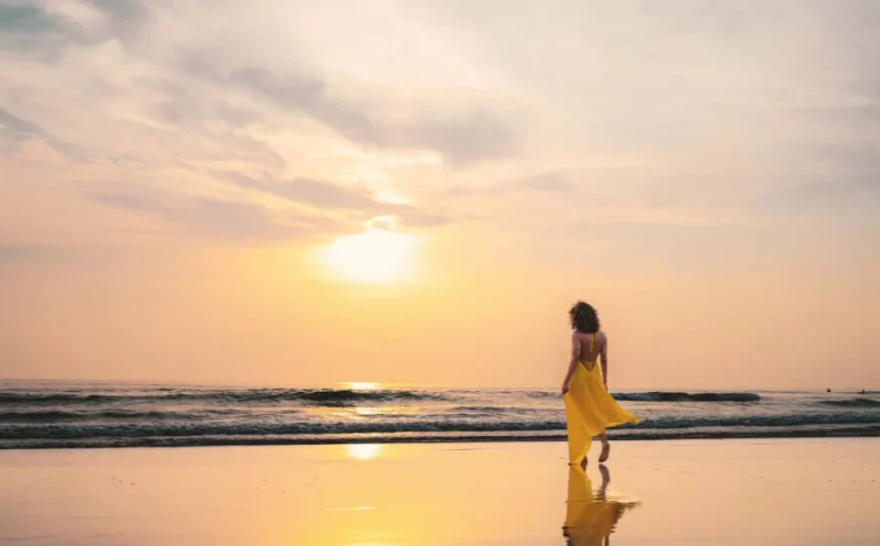 A woman in a flowing yellow dress walks along a calm beach at golden hour, her reflection mirrored in the wet sand. The soft pastel sky and gentle waves capture the essence of slow travel — taking time to be present, unhurried, and fully immersed in the beauty of a destination rather than rushing through it.