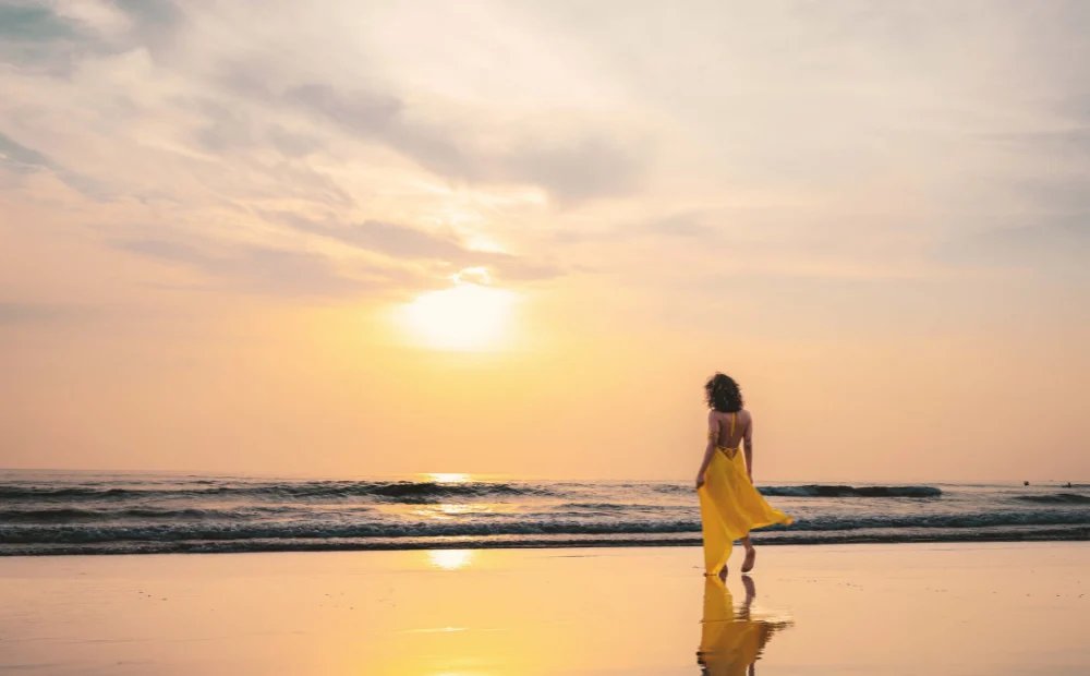A woman in a flowing yellow dress walks along a calm beach at golden hour, her reflection mirrored in the wet sand. The soft pastel sky and gentle waves capture the essence of slow travel — taking time to be present, unhurried, and fully immersed in the beauty of a destination rather than rushing through it.