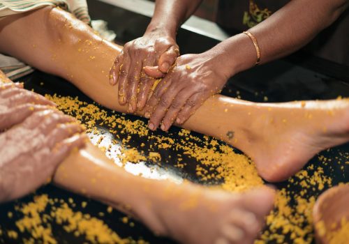 Ayurvedic therapist performing Udvartana therapy using a herbal powder mixture on a guest's legs during a traditional Ayurvedic treatment session.