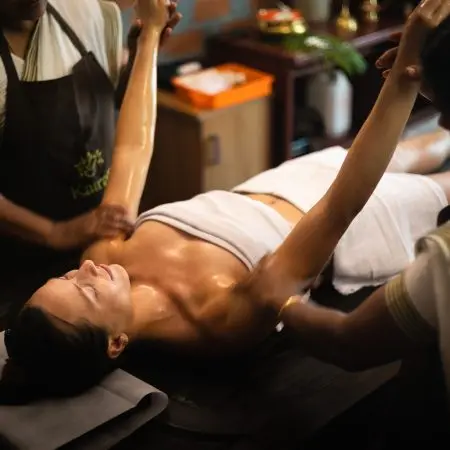 Two therapists performing Ayurvedic postpartum care oil massage on a woman lying on a treatment table