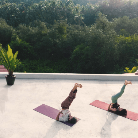 Two people practicing shoulder stand yoga poses on a rooftop terrace surrounded by tropical greenery