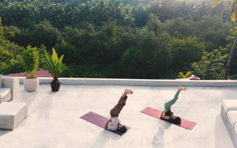 Two people practicing shoulder stand yoga poses on a rooftop terrace surrounded by tropical greenery