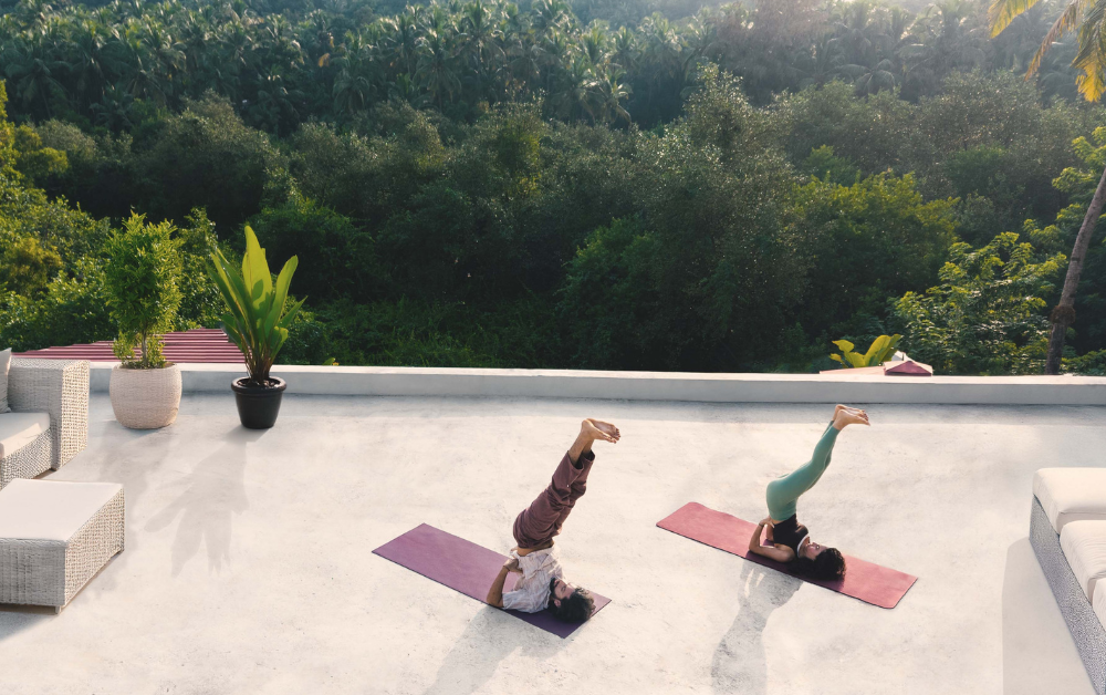 Two people practicing shoulder stand yoga poses on a rooftop terrace surrounded by tropical greenery