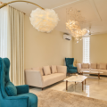 Elegant living room with teal wingback chairs, cream sofas, and a gold arc floor lamp beneath a feather pendant light.