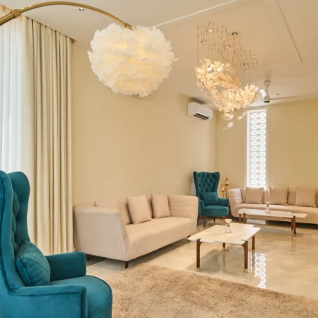 Elegant living room with teal wingback chairs, cream sofas, and a gold arc floor lamp beneath a feather pendant light.