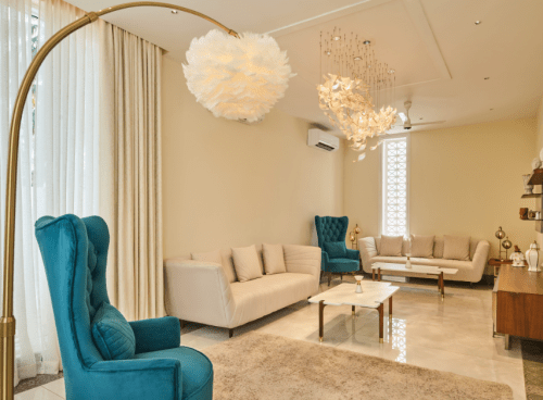 Elegant living room with teal wingback chairs, cream sofas, and a gold arc floor lamp beneath a feather pendant light.