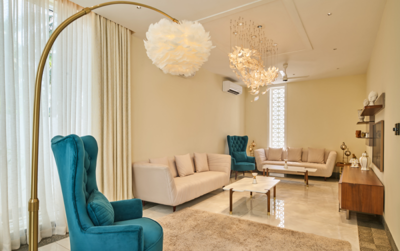 Elegant living room with teal wingback chairs, cream sofas, and a gold arc floor lamp beneath a feather pendant light.